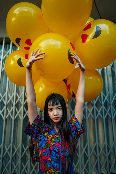 an Asian girl in bright clothes posing for a picture with big yellow smiley face balloons